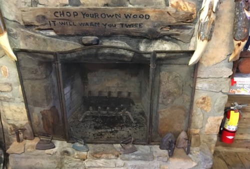 A rustic stone fireplace with a wooden sign above that reads, "CHOP YOUR OWN WOOD IT WILL WARM YOU TWICE."