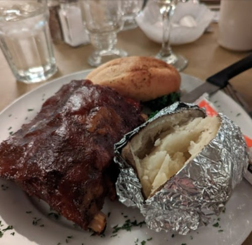 A plate with BBQ ribs, a baked potato wrapped in foil, and a bread roll, served on a white dish with garnish.