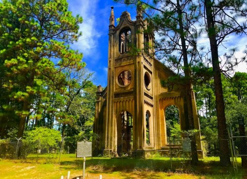 A tall, weathered stone structure surrounded by trees, with arched windows and a bell tower, set in a lush green landscape.