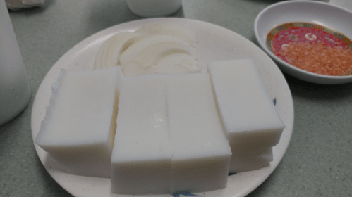 A plate with sliced white tofu and a side of dipping sauce with garnishes.