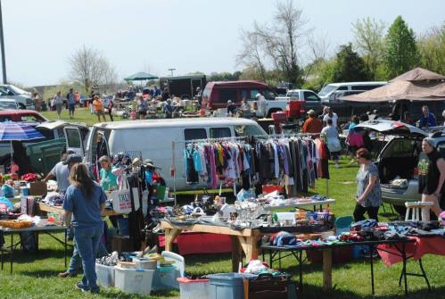 A bustling outdoor flea market with vendors, clothing racks, and shoppers browsing various items on tables.