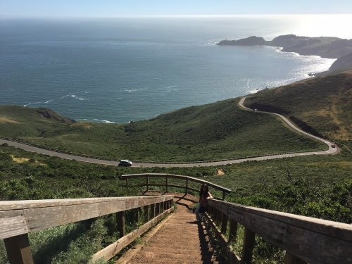 A scenic view from a wooden staircase overlooking a coastline and winding road, with lush green hills and ocean.