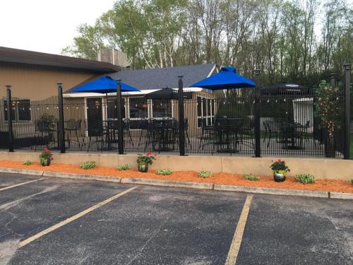 Outdoor patio area with black fencing, blue umbrellas, and flower beds, adjacent to a building surrounded by trees.