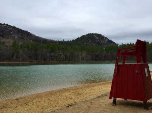 A red lifeguard chair on a sandy beach by a calm lake, surrounded by green trees and mountains under a cloudy sky.