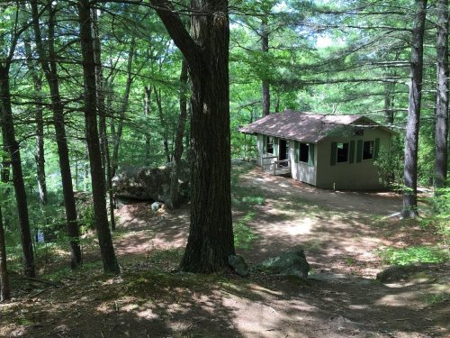 A small cabin nestled among green trees, with a rocky area nearby and a path leading to it.