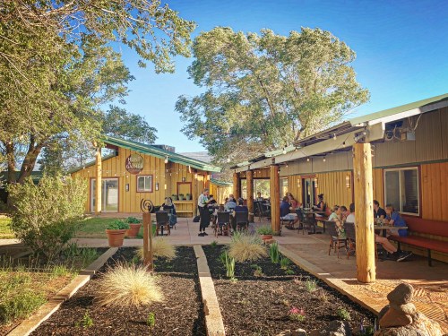 A sunny outdoor dining area with people seated at tables, surrounded by greenery and rustic buildings.