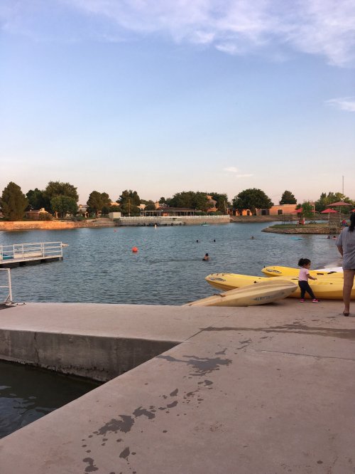 A calm lake scene with people swimming and kayaks on the shore, surrounded by trees and a clear sky.