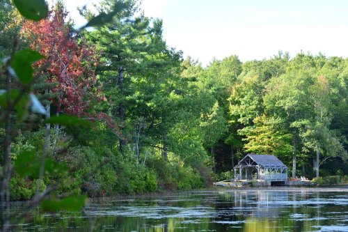 A serene lakeside scene with a wooden gazebo surrounded by lush green trees and hints of autumn colors.