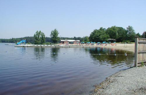 A serene lakeside scene with a sandy beach, swimming area, and a water slide under a clear blue sky.