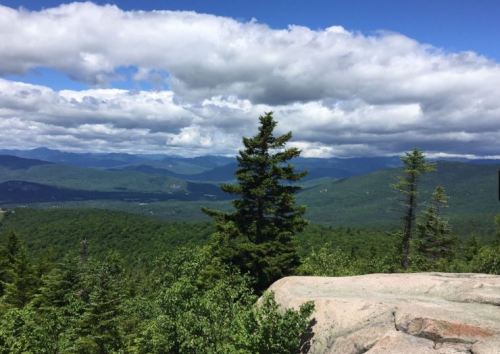 A scenic view from a mountain peak, featuring lush green forests and distant mountains under a partly cloudy sky.