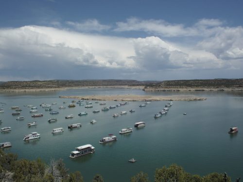 A serene lake scene with numerous houseboats anchored, surrounded by hills and a cloudy sky.