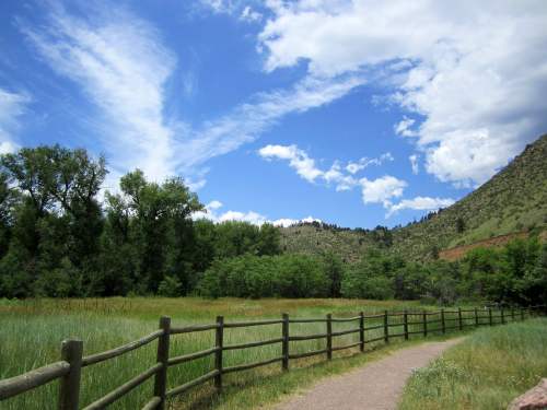 A scenic path winds through a green field, bordered by a wooden fence, under a blue sky with fluffy clouds.