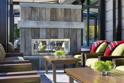 Cozy outdoor seating area with a modern fireplace, green cushions, and decorative plants under a shaded patio.