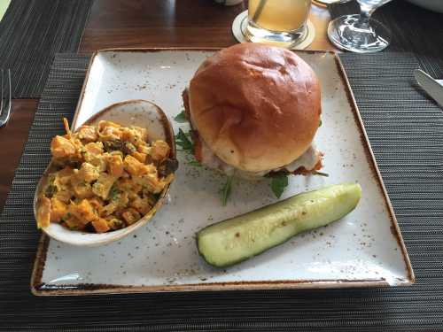 A plate with a sandwich, a side of creamy corn salad, and a cucumber slice, set on a textured tablecloth.