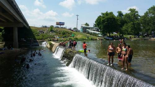 People enjoying a sunny day by a waterfall, with some swimming and others relaxing by the water's edge.