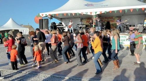 A lively crowd dances at a festival stage with a band, surrounded by festive decorations and a sunny atmosphere.