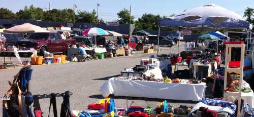 A bustling flea market with various stalls, tents, and items for sale, set in a parking lot on a sunny day.