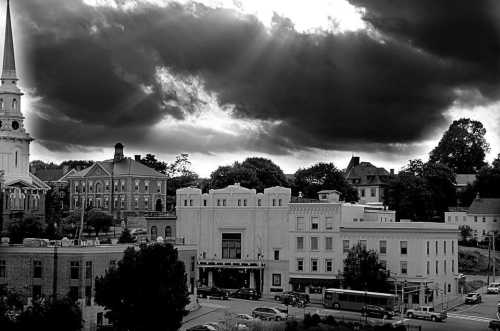 A black and white cityscape featuring buildings, a church steeple, and dramatic clouds overhead.