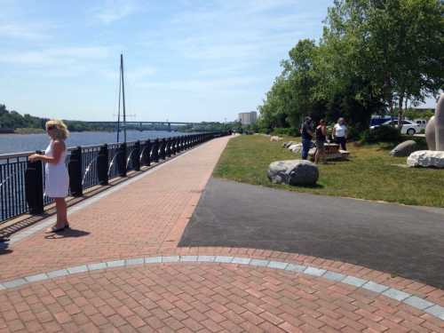 A scenic riverside path with people enjoying the outdoors, greenery, and a bridge in the background under a clear blue sky.