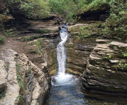 A serene waterfall cascades through rocky terrain, surrounded by lush greenery and steep cliffs.