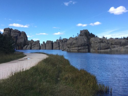 A serene landscape featuring a winding path beside a calm lake, surrounded by towering rock formations and blue skies.