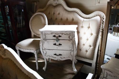 A vintage white nightstand with three drawers, placed beside an upholstered chair and a tufted headboard.
