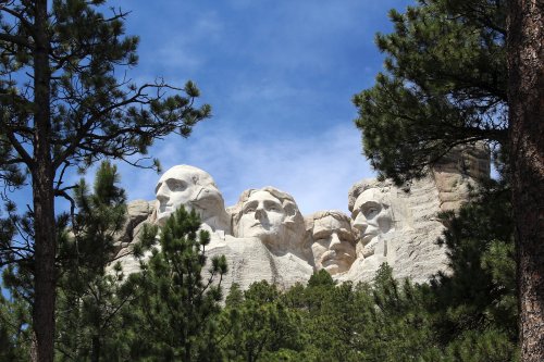 Mount Rushmore, featuring four presidential faces, framed by green trees and a blue sky.