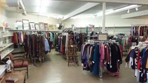 A spacious thrift store interior with racks of clothing, framed pictures on the walls, and a chair in the corner.