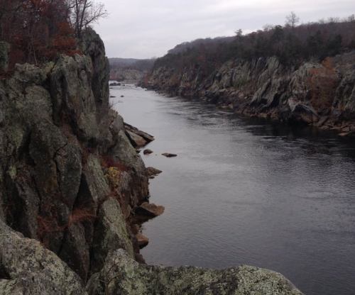 A serene river flows between rocky cliffs, surrounded by trees under a cloudy sky.