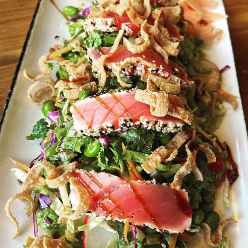 A vibrant salad topped with seared tuna, crispy toppings, and a variety of fresh vegetables, drizzled with sauce.