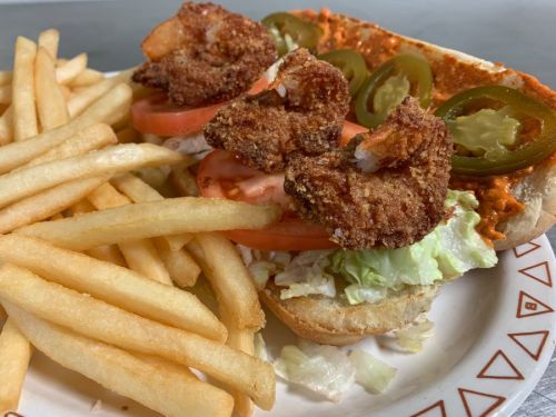 A plate with a shrimp sandwich topped with lettuce, tomatoes, jalapeños, and fries on the side.
