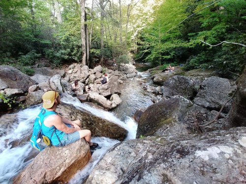 A person sits on a rock by a flowing stream in a lush forest, with others relaxing nearby on the rocks.