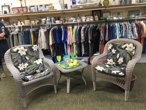 Two wicker chairs with floral cushions and a small table with drinks and fruit, set against a backdrop of hanging clothes.