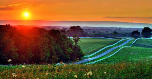 A scenic landscape at sunset, featuring rolling hills, a winding road, and vibrant colors in the sky.