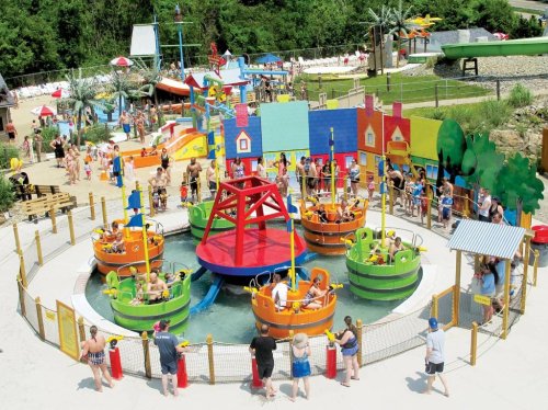 A colorful water park area with boats, children playing, and adults watching from the sidelines on a sunny day.