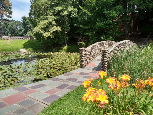 A stone bridge arches over a pond filled with lily pads, surrounded by lush greenery and vibrant orange flowers.