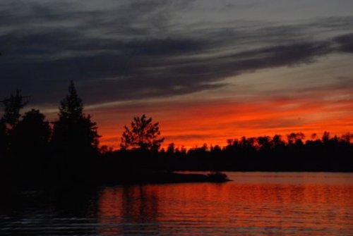 A serene lake at sunset, with vibrant orange and red hues reflecting on the water and silhouetted trees in the foreground.