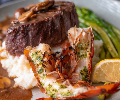A gourmet plate featuring steak, grilled lobster, asparagus, and lemon, served over creamy mashed potatoes.