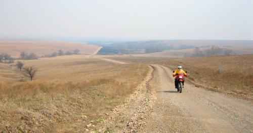 A motorcyclist rides along a winding dirt road through a vast, open landscape with rolling hills and sparse trees.