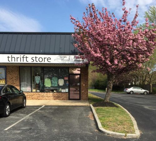 A thrift store with a sign, featuring a blooming pink tree in front and parked cars in the lot.