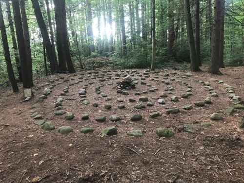 A stone labyrinth in a forest, surrounded by trees, with a small fire pit at the center.