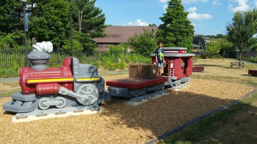 A child stands on a playground train structure made of colorful plastic, surrounded by gravel and greenery.
