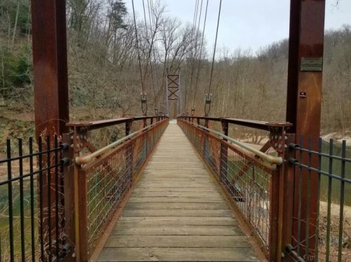 A wooden suspension bridge stretches over a calm river, surrounded by bare trees and a cloudy sky.