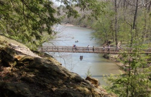 A scenic view of a river with a wooden bridge and people walking, surrounded by lush greenery and rocky terrain.
