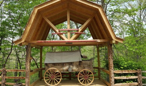A wooden shelter with a wagon underneath, surrounded by lush green trees.