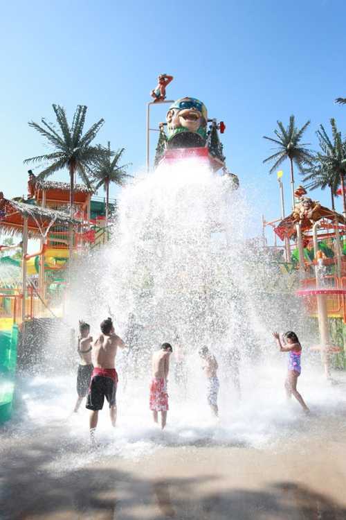 Children play in a water park, splashing under a large water feature with palm trees in the background.