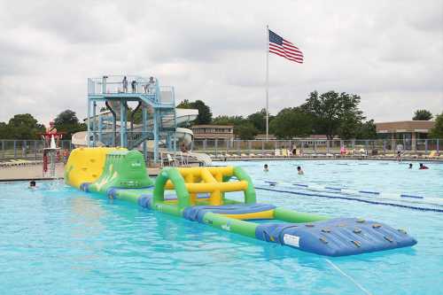 A colorful inflatable water slide and obstacle course at a public pool, with swimmers and a lifeguard on duty.