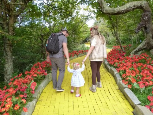 A family walks hand-in-hand along a yellow brick path surrounded by vibrant red flowers and lush greenery.