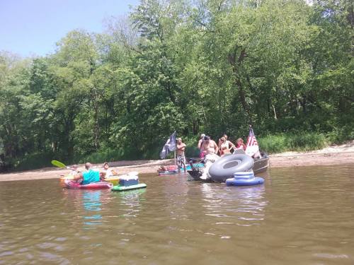 A group of people on inflatable rafts and a boat enjoying a sunny day on a river surrounded by trees.