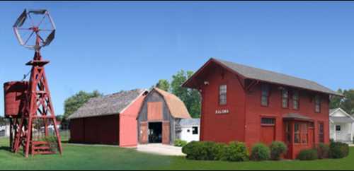 A red barn and a two-story red house with a windmill nearby, set against a clear blue sky.